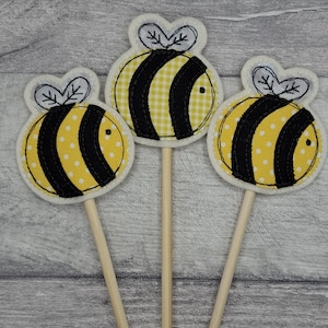 May include: Three bee-shaped decorations in yellow and black, mounted on wooden sticks. The bees feature black stripes and either white polka dots or a yellow gingham pattern. Each has a white outline and a white and black wing design.