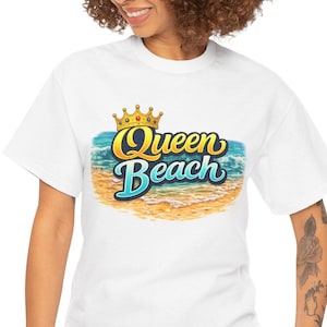 Può includere: T-shirt bianca con la scritta "Queen Beach" in un design colorato, che include una corona, onde dell'oceano e una spiaggia sabbiosa. La maglietta è realizzata in un materiale morbido.