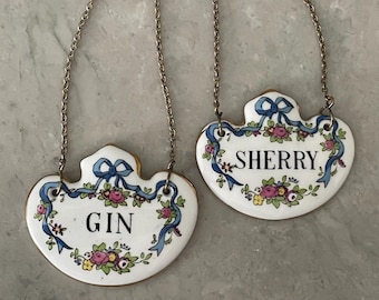 May include: Two heart-shaped ceramic liquor labels with silver chains. One reads "GIN" and the other "SHERRY" in black text. Both are white with blue ribbons and floral designs, and gold trim.