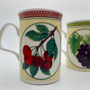 May include: Two ceramic mugs with a yellow background and a white circle in the centre. The mug on the left has a red cherry branch with green leaves. The mug on the right has a purple grape vine with green leaves.