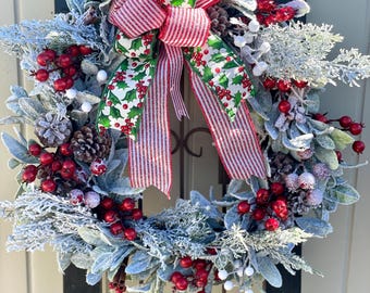 May include: A festive Christmas wreath with frosted greenery, red berries, and pinecones. The wreath features a large bow with a holly and stripe pattern. The wreath is mounted on a dark green rectangular frame.