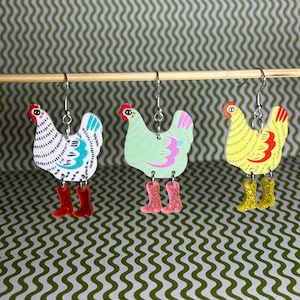 May include: Three pairs of colourful chicken earrings. Each pair features a different colour and pattern: white with blue accents, light green with pink accents, and yellow with red accents. The earrings are made of wood and have a whimsical, folk art style.