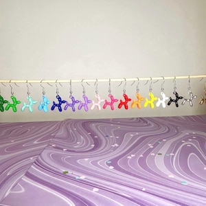 Può includere: Un set di 14 orecchini a forma di cane di palloncino in vari colori, tra cui verde, blu, viola, rosa, arancione, giallo, nero, argento e oro. Gli orecchini sono appesi a un'asta argentata.