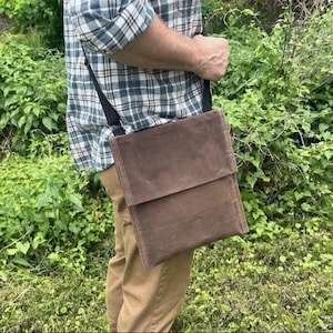 May include: A brown waxed canvas crossbody bag with a rectangular flap closure and a black adjustable shoulder strap. The bag features a smooth, matte finish and a structured, flat profile, designed to be worn across the body for carrying items.