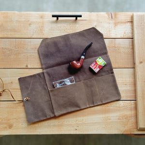 May include: A brown canvas tobacco pouch with multiple pockets, holding a wooden pipe, a small pack of cigarettes, and a clear plastic container. The pouch is open, revealing its contents. The items are on a wooden surface.