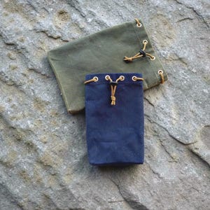 May include: Two rectangular drawstring pouches made of heavy canvas fabric. One pouch is olive green and the other is navy blue. Each bag features metal grommets along the top edge, a tan cord drawstring, and a small black plastic cord lock.