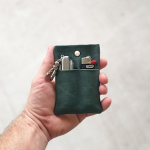 May include: A dark green canvas lighter case with a snap closure. The case holds a silver lighter and a black and yellow BIC lighter. A small lanyard with a patterned knot hangs from the case.