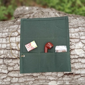 May include: A dark green canvas roll-up organizer featuring three interior pockets. The pockets contain a small rectangular box of matches, a polished brown wooden tobacco pipe, and a clear plastic pouch filled with loose tobacco.