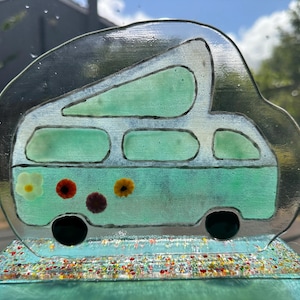 May include: A decorative glass camper van with a light teal body and black wheels. The van is adorned with colorful flowers and sits atop a glittery base. The image is set against a blurred outdoor background.