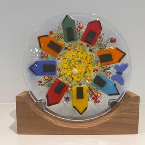 May include: A decorative glass plate with a colourful design. The plate features a central yellow area surrounded by colourful shapes, including red, orange, green, and blue. It is displayed on a wooden stand.