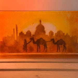 以下が含まれることがあります： 砂漠のシーンを描いた長方形のガラスアート作品。このアートワークは、都市のスカイラインのシルエット、2頭のラクダ、そしてそれらを導く人物を特徴としており、すべて茶色と黒の色合いで、オレンジと黄色の夕日の背景に描かれています。