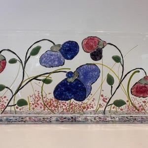 Puede incluir: Una pieza de arte en vidrio rectangular con un diseño floral. La obra presenta flores estilizadas en tonos azules y rojos, con hojas verdes y delicados tallos. La base es un mosaico de piezas de vidrio de colores.