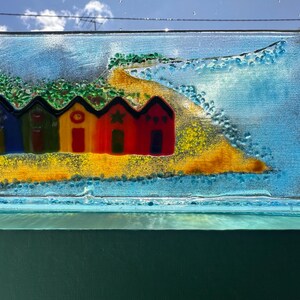 Könnte beinhalten: Ein rechteckiges Glaskunstwerk, das eine Strandszene darstellt. Fünf bunte Strandhütten stehen an einem gelben Sandstrand, mit einem blauen Ozean- und Himmelshintergrund. Die Hütten sind in Blau, Grün, Gelb, Rot und Orange bemalt.
