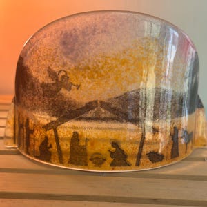 Op de afbeelding: Een keramisch, koepelvormig decoratief object met een geschilderde kerststal. De scène toont silhouetten van figuren, een stal en een engel tegen een achtergrond van gele, bruine en paarse tinten.