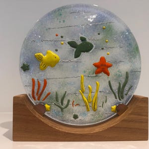 Peut inclure: Une assiette décorative en verre avec une scène sous-marine, représentant un poisson jaune, un poisson vert et une étoile de mer orange. L'assiette est soutenue par un support en bois. Le design comprend des plantes marines et des bulles, créant un thème marin.