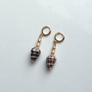 May include: A pair of gold-colored hoop earrings with a dangling design. Each earring features a small, cone-shaped shell with a brown and white pattern, accented by a string of small, light-colored beads. The earrings are set against a white background.