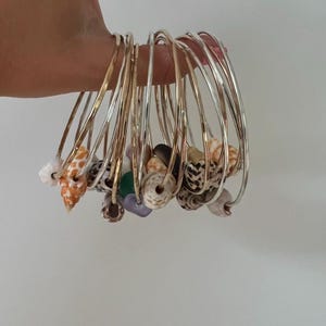 May include: A set of thin, metallic bangles in gold and silver tones, each featuring a single small seashell or bead charm. The charms include various patterns and colors, such as spotted, orange, white, and purple, threaded onto the metal bands.