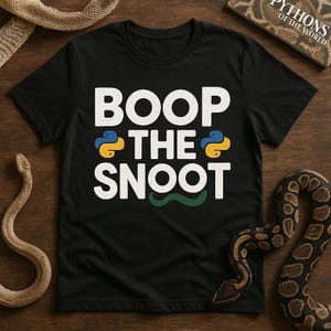 May include: Black t-shirt with the text "BOOP THE SNOOT" in white, with a snake graphic. The shirt is on a wooden surface with snakes and a book titled "Pythons of the World".