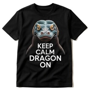 May include: Black t-shirt featuring a detailed illustration of a dragon's face in shades of blue, gray, and brown. The text "KEEP CALM DRAGON ON" is printed in white below the dragon's image. The shirt has a crew neck and short sleeves.