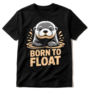 May include: Black t-shirt featuring a cartoon otter in water with the text "BORN TO FLOAT" in a bold, playful font. The otter is gray and white with a light brown snout, and the water is a light brown color.