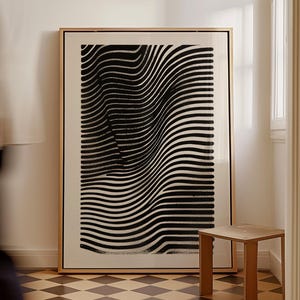 May include: A black and white abstract print in a wooden frame hangs on a white wall above a small wooden stool. The print features a series of curved lines that create a sense of movement and depth.