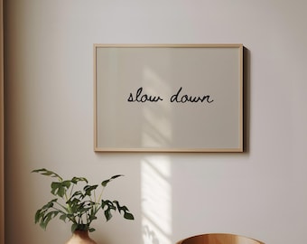 May include: A framed print with the words "slow down" in black script. The print is displayed on a neutral-colored wall, with a wooden chair and a small table with a vase of greenery in the foreground. The room is bathed in natural light.