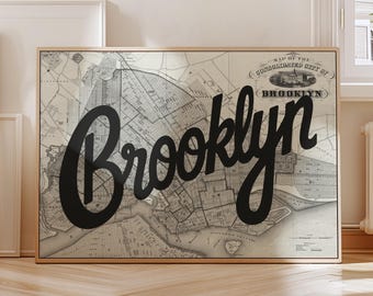 May include: A framed map print of Brooklyn, New York, with the word "Brooklyn" in large, black cursive script. The map is in grayscale, showing streets and buildings. The frame is a light wood color.