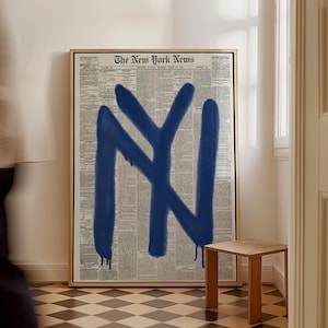 May include: A framed artwork featuring a vintage newspaper background with a large, blue graffiti-style "NY" symbol. The artwork is displayed in a room with a small wooden side table and a checkered floor.