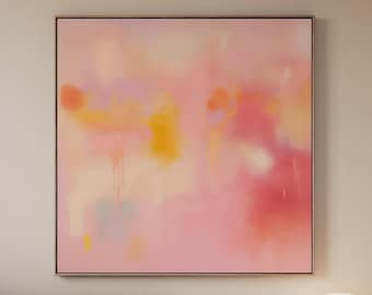 May include: Abstract art print with a soft, blurred composition in shades of pink, yellow, and orange. The artwork is framed in a thin, dark frame and hangs above a beige sofa and wooden table.