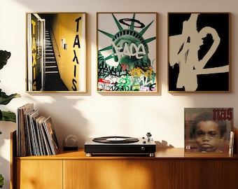 May include: Three framed art prints with gold frames hang above a wooden cabinet. The prints feature a yellow taxi door, the Statue of Liberty with graffiti, and an abstract design. A record player and vinyl records sit on the cabinet.