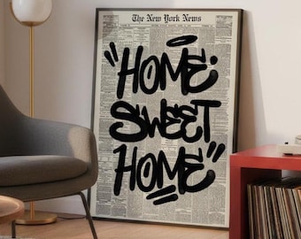 May include: A framed black and white print with the words "HOME SWEET HOME" in bold black graffiti-style lettering. The background is a newspaper print. The print is displayed in a living room setting.