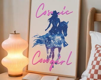 May include: A framed art print featuring a blue silhouette of a cowgirl riding a horse, with the words "Cosmic Cowgirl" in pink script. The print is displayed on a nightstand next to a lamp and an open book.
