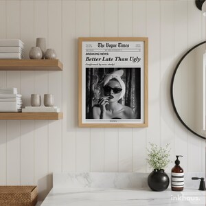 May include: A framed black and white wall art print titled The Vogue Times with the headline Breaking News: Better Late Than Ugly. The image features a person wearing a towel wrap on their head and dark sunglasses, applying lipstick.