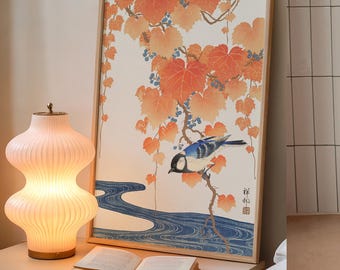 May include: A framed art print featuring a blue and white bird perched on a branch with orange leaves and blue berries. A decorative lamp with a ribbed design sits next to an open book. The artwork has a light wood frame.