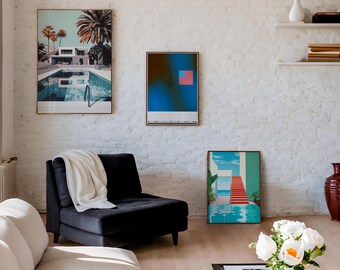 May include: Three framed art prints on a white brick wall. The prints feature abstract designs and a scene of a house with a pool. A black armchair with a white throw blanket sits in front of the wall. A vase of white flowers sits on a coffee table in the foreground.