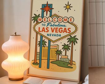 May include: A framed print featuring the iconic "Welcome to Fabulous Las Vegas Nevada" sign. The artwork includes palm trees, stars, and a casino, with a warm color palette. A decorative lamp and an open book are on a nearby table.