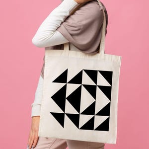 May include: A cream-colored tote bag featuring a geometric design of black triangles. The bag is held against a pink background. The design is a repeating pattern of triangles.