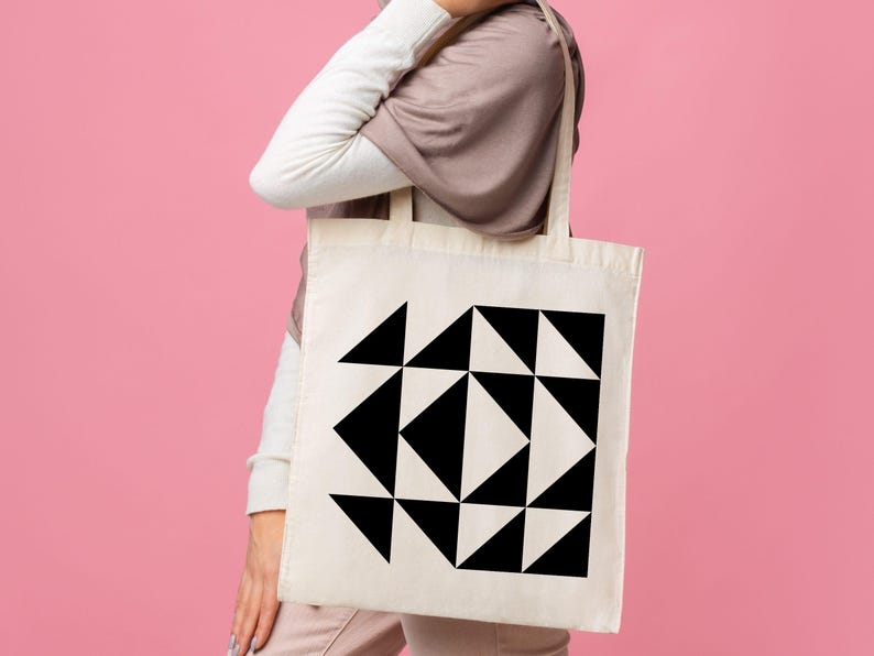 May include: A cream-colored tote bag featuring a geometric design of black triangles. The bag is held against a pink background. The design is a repeating pattern of triangles.