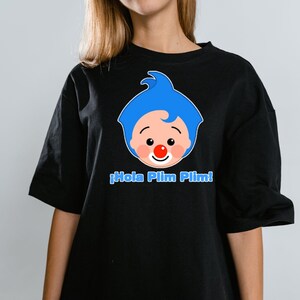 May include: Black t-shirt featuring a cartoon clown face with blue hair, a red nose, and the text "&iexcl;Hola Plim Plim!" in blue. The shirt is a casual style, suitable for everyday wear.