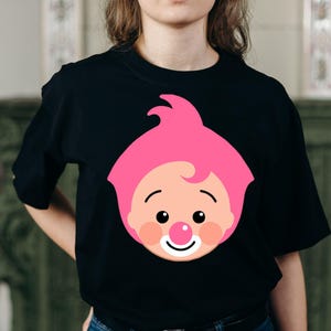May include: Black t-shirt featuring a cartoon face with pink hair, rosy cheeks, a red nose, and a smiling mouth. The graphic is centered on the front of the shirt. The shirt is a crew neck style.