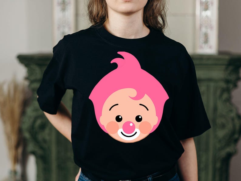 May include: Black t-shirt featuring a cartoon face with pink hair, rosy cheeks, a red nose, and a smiling mouth. The graphic is centered on the front of the shirt. The shirt is a crew neck style.