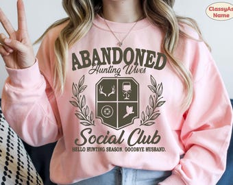 Op de afbeelding: Een lichtroze sweatshirt met de tekst "ABANDONED Hunting Wives Social Club" in donkergroen. Het ontwerp bevat een wapenschild met geweien, een wijnglas, een doel en een vogel. De zin "Hello Hunting Season, Goodbye Husband" staat eronder.