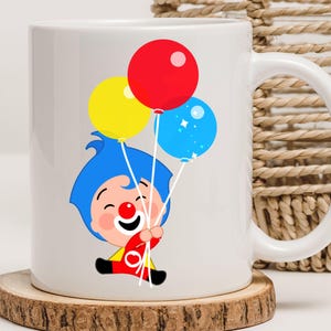 May include: White ceramic mug featuring a cartoon illustration of a clown holding three balloons. The balloons are red, yellow, and blue. The clown has blue hair and is wearing a red and yellow outfit. The mug sits on a wooden coaster.