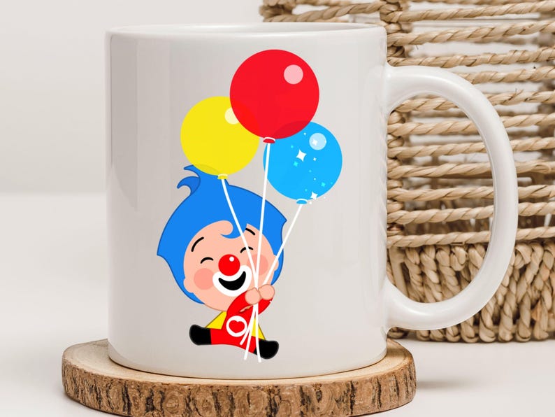 May include: White ceramic mug featuring a cartoon illustration of a clown holding three balloons. The balloons are red, yellow, and blue. The clown has blue hair and is wearing a red and yellow outfit. The mug sits on a wooden coaster.