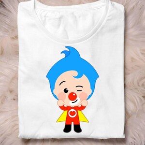 May include: White t-shirt featuring a cartoon character with blue hair, a red nose, and a red and yellow outfit. The character is winking and has its arms raised. The shirt is folded on a white surface.