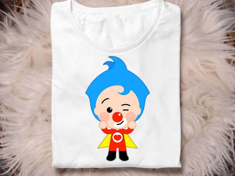 May include: White t-shirt featuring a cartoon character with blue hair, a red nose, and a red and yellow outfit. The character is winking and has its arms raised. The shirt is folded on a white surface.