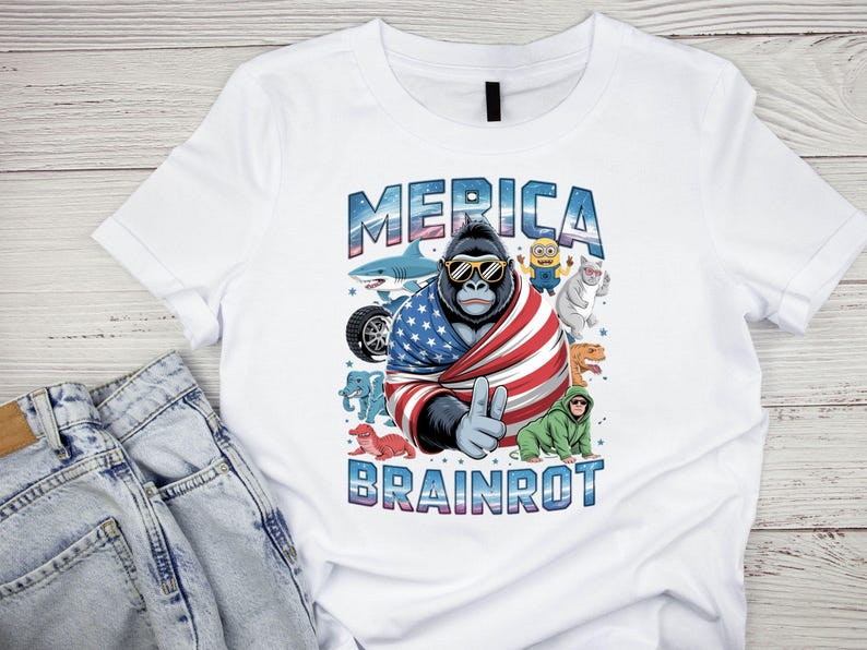 Może przedstawiać: Biała koszulka z gorylem w okularach przeciwsłonecznych i amerykańską flagą, z napisem "MERICA" powyżej i "BRAINROT" poniżej. Zawiera kresk&oacute;wkowe obrazy rekina, Minionka, kota, dinozaura i innych.
