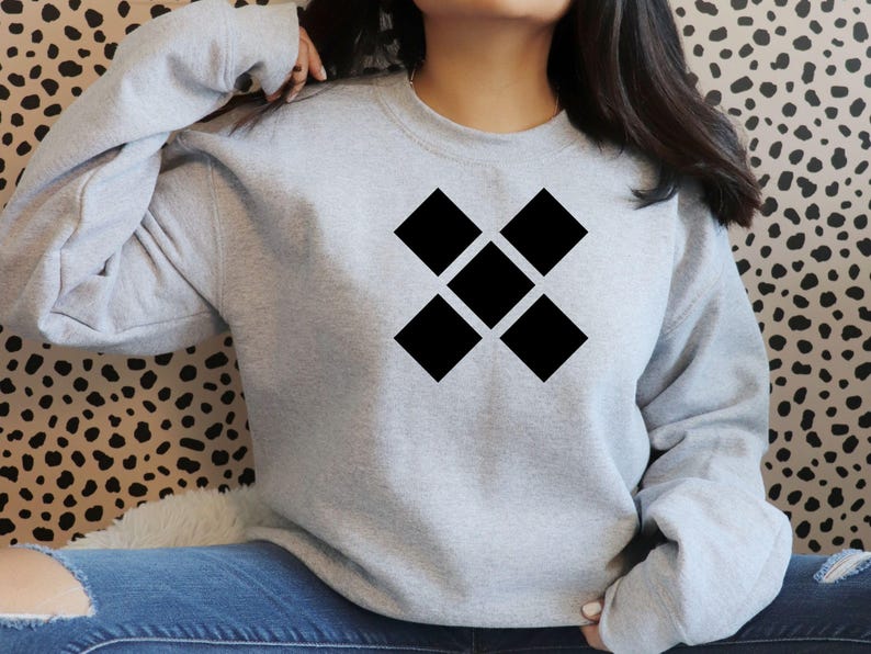 May include: A light gray sweatshirt featuring a black geometric design of five connected diamonds. The sweatshirt has a crew neck and long sleeves. The background is a spotted pattern.