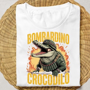 Może przedstawiać: Biała koszulka z kresk&oacute;wkowym aligatorem w mundurze wojskowym, ze słowami "BOMBARDINO CROCODILO" w kolorze ż&oacute;łtym. Aligator ma otwartą paszczę i jest umieszczony na pomarańczowo-ż&oacute;łtej eksplozji.