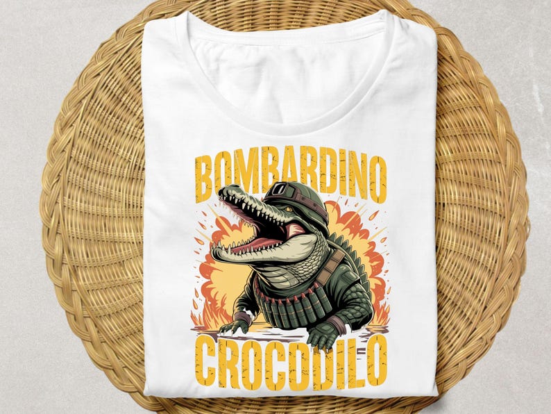 Może przedstawiać: Biała koszulka z kresk&oacute;wkowym aligatorem w mundurze wojskowym, ze słowami "BOMBARDINO CROCODILO" w kolorze ż&oacute;łtym. Aligator ma otwartą paszczę i jest umieszczony na pomarańczowo-ż&oacute;łtej eksplozji.